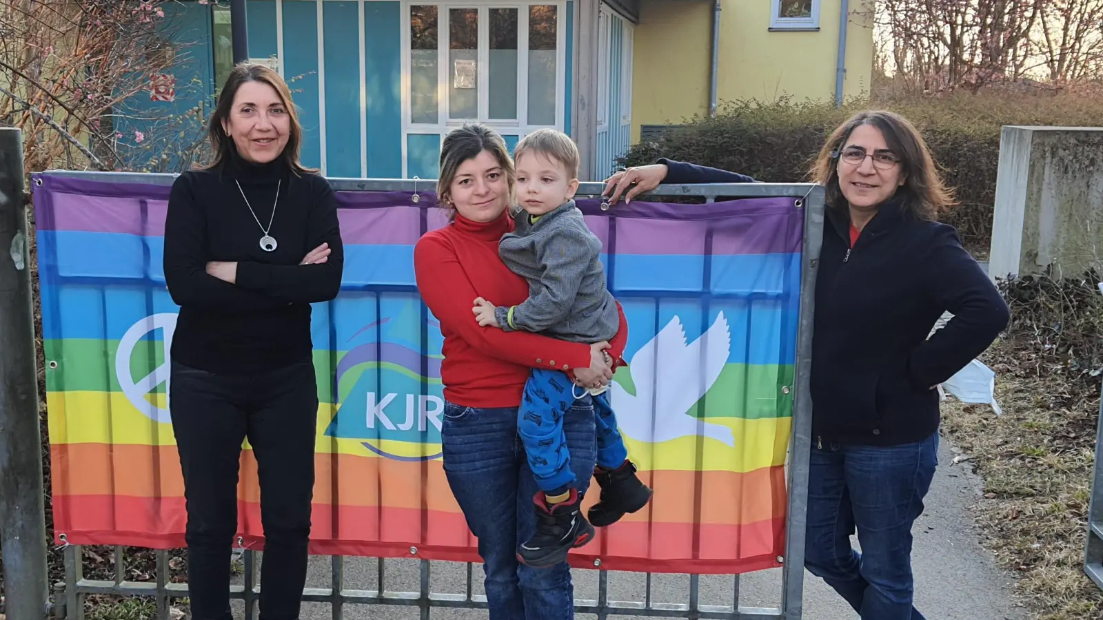 Yuliia Zadyraka ist mit ihrem vierjährigen Sohn Maksym vor dem Raketenbeschuss in Charkiw geflohen. Jetzt wohnt sie bei ihrer früheren Chefin Züleyha Yilmaz (rechts), der Leiterin des Jugendtreff Neuaubing. Auch die Familie von Zadyrakas Cousin ist geflüchtet und bei der Mutter von Yilmaz‘ Kollegin Johanna Baindl untergekommen (links). Im Hintergrund das KJR-Friedensbanner, das derzeit an vielen Freizeitstätten in München hängt. (Foto: KJR München-Stadt)